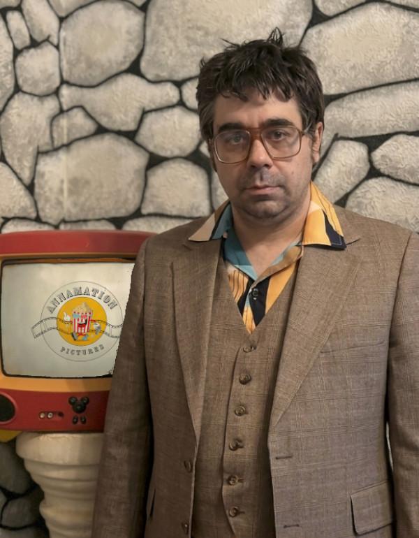 photo of the male director wearing a brown suit standing next to a TV set showing his film logo