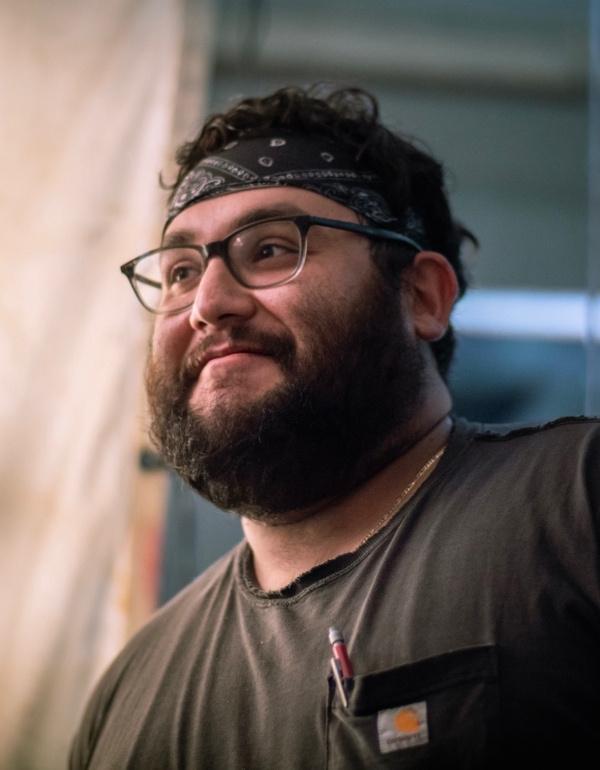 A smiling bearded man wearing glasses and a black bandana looks off-camera in a candid close-up.