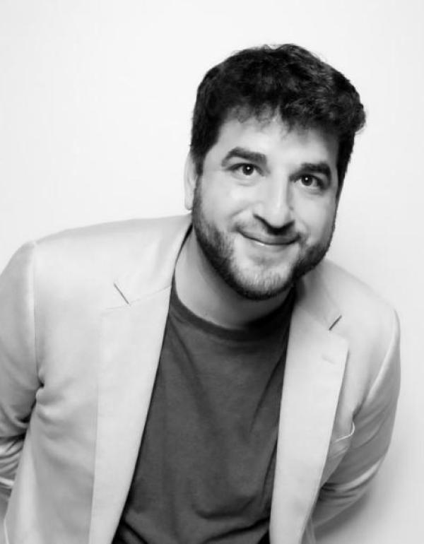 director, Zach Wolff in a suit jacket and shirt smiles at the camera in black and white