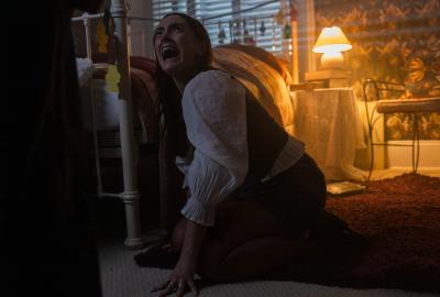 A woman on her hands and knees in a dark bedroom, screaming at a figure standing in front of her.