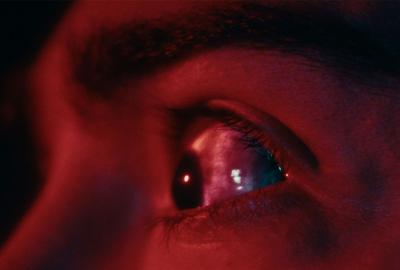 Close-up of a human eye lit in red tones, looking with reflections of light visible on the pupil.