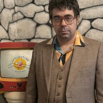 photo of the male director wearing a brown suit standing next to a TV set showing his film logo