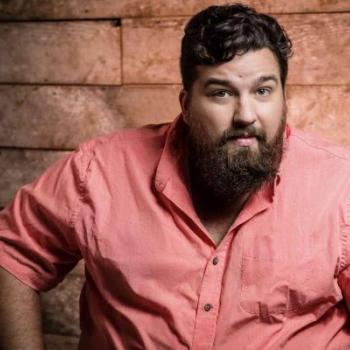 A handsome man poses in front of a wooden wall wearing a pink shirt