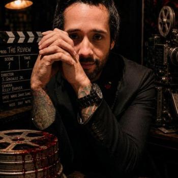 Director Samuel Gonzalez Jr. in a cinematic portrait surrounded by vintage filmmaking equipment and blood-streaked film reels, representing his horror film THE REVIEW.