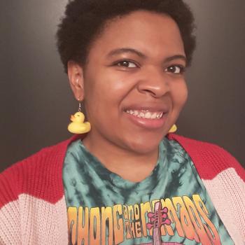 Woman with short hair in a pink sweater with rubber duck earrings smiling.
